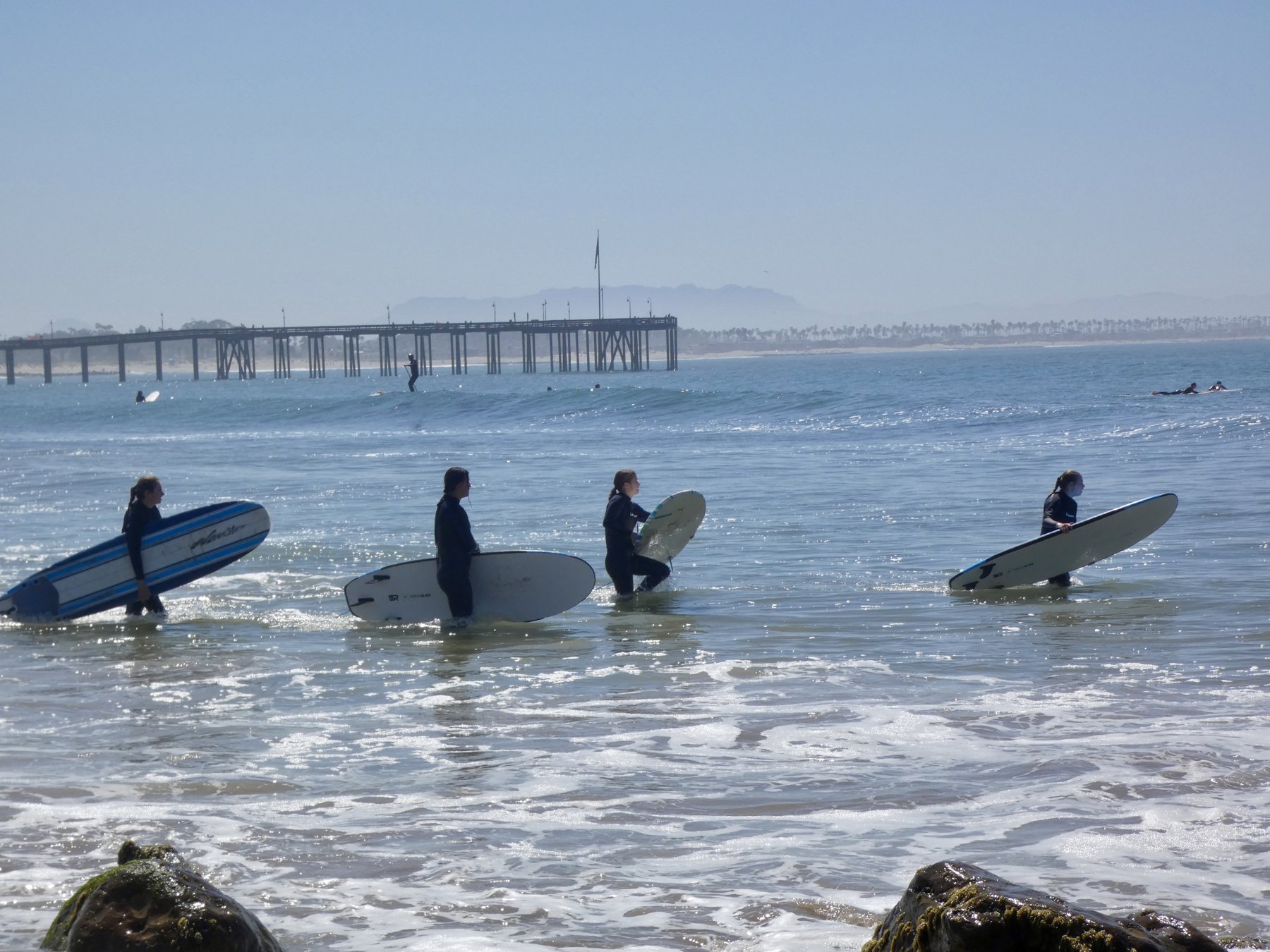 Surfers wading out with longboards near a pier