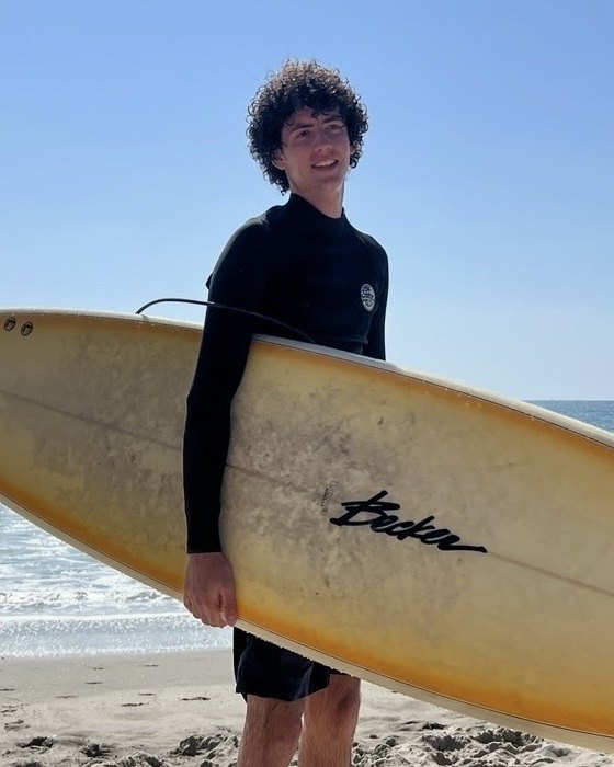Max Plush on the beach in a wetsuit, holding a yellow surfboard