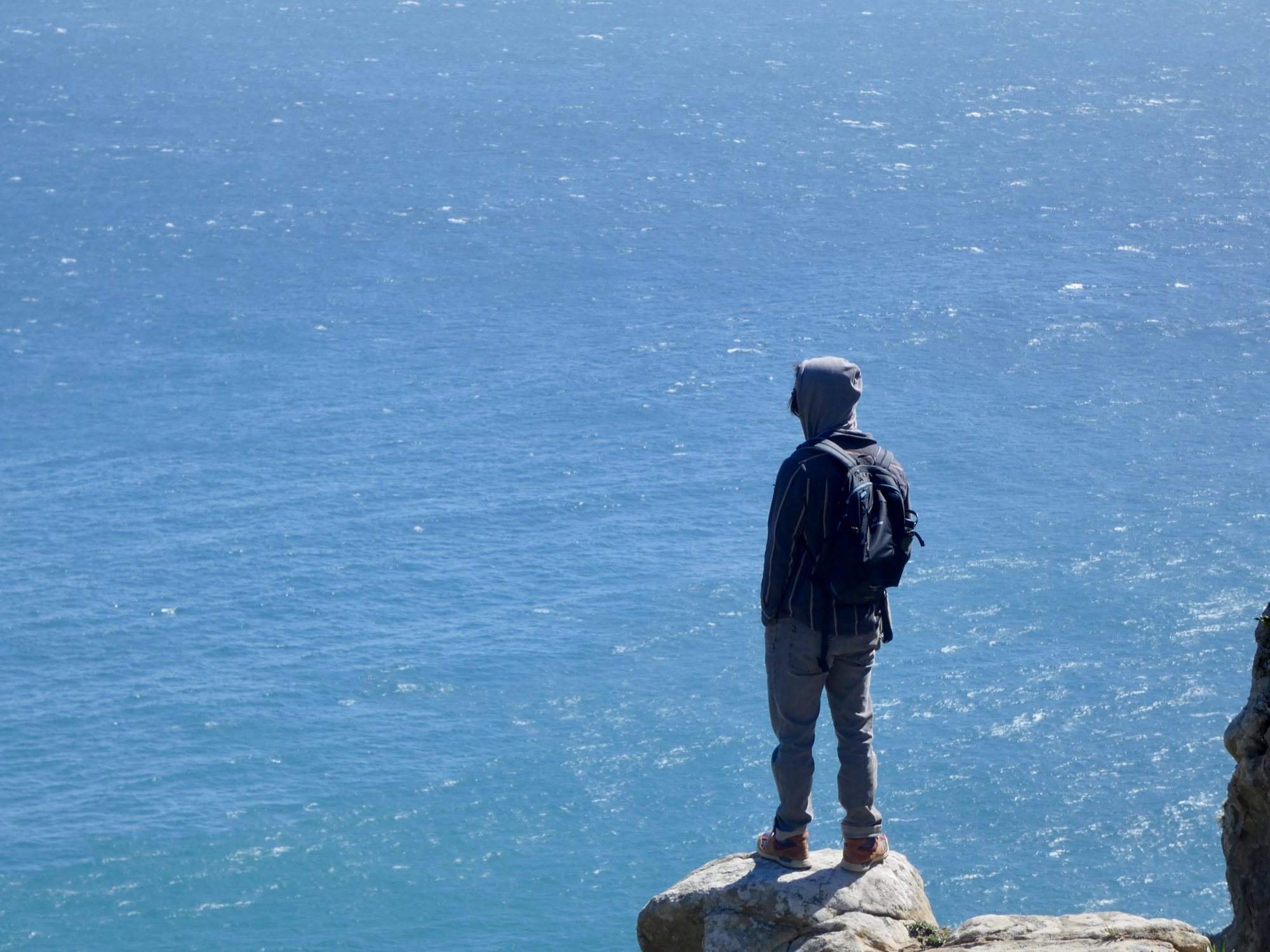 Hooded figure standing on a rock over open blue ocean