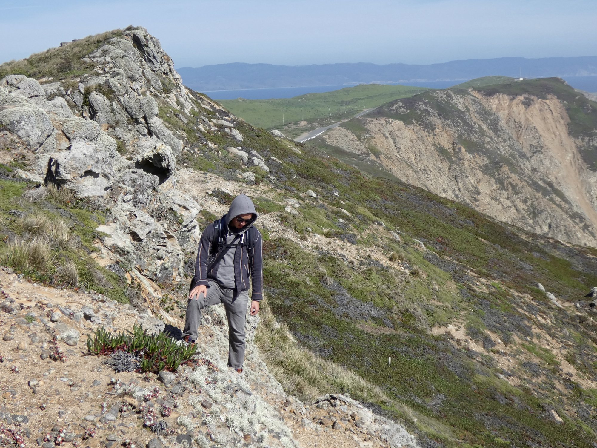 Hiker in a hood climbing a rocky coastal ridge above green headlands